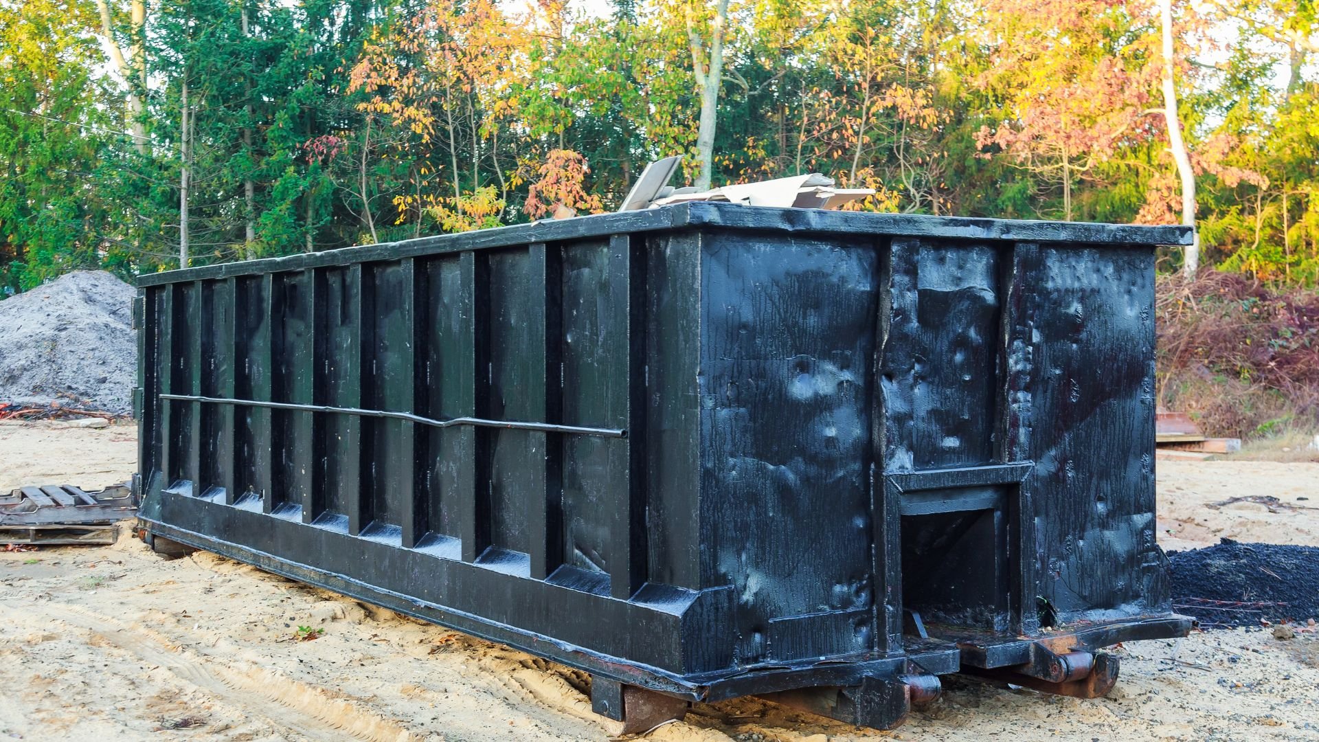 Large blue-gray dumpster on sandy ground with autumn trees in background